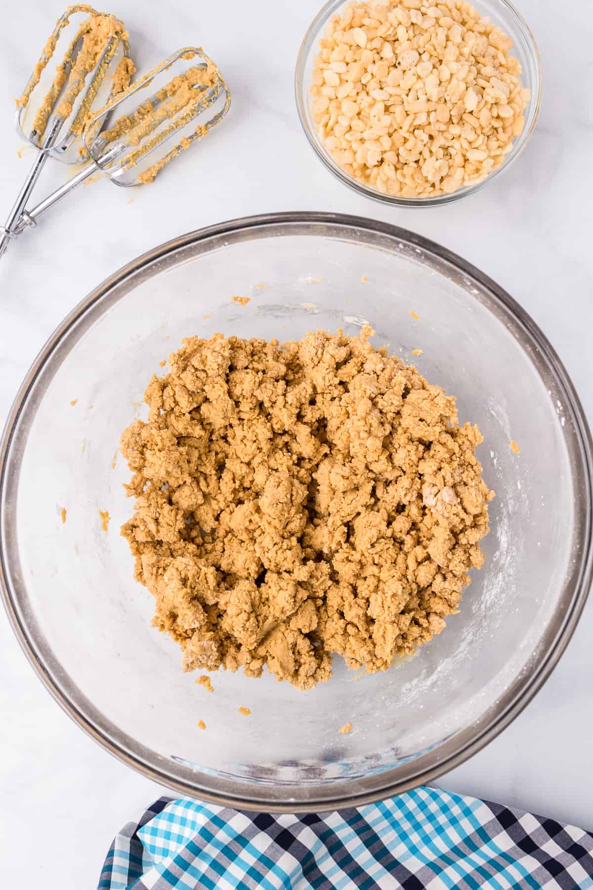 Chunky peanut butter dough mixed in a large bowl with a bowl from overhead on a counter with a bowl of rice krispies cereal and peanut butter covered mixer beaters on the counter nearby.