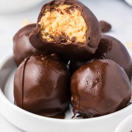 Peanut butter balls with rice krispies stacked on a small plate on a counter from the side with the top truffle missing a bite to see the inside.