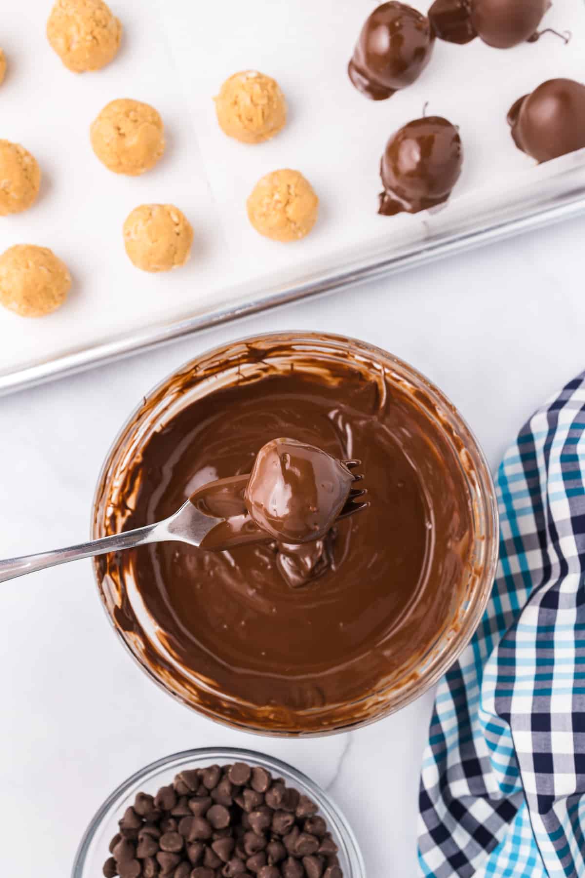 Peanut butter balls with rice krispies being dipped into a bowl of chocolate from overhead with a fork with more peanut butter balls on a cookie sheet nearby.
