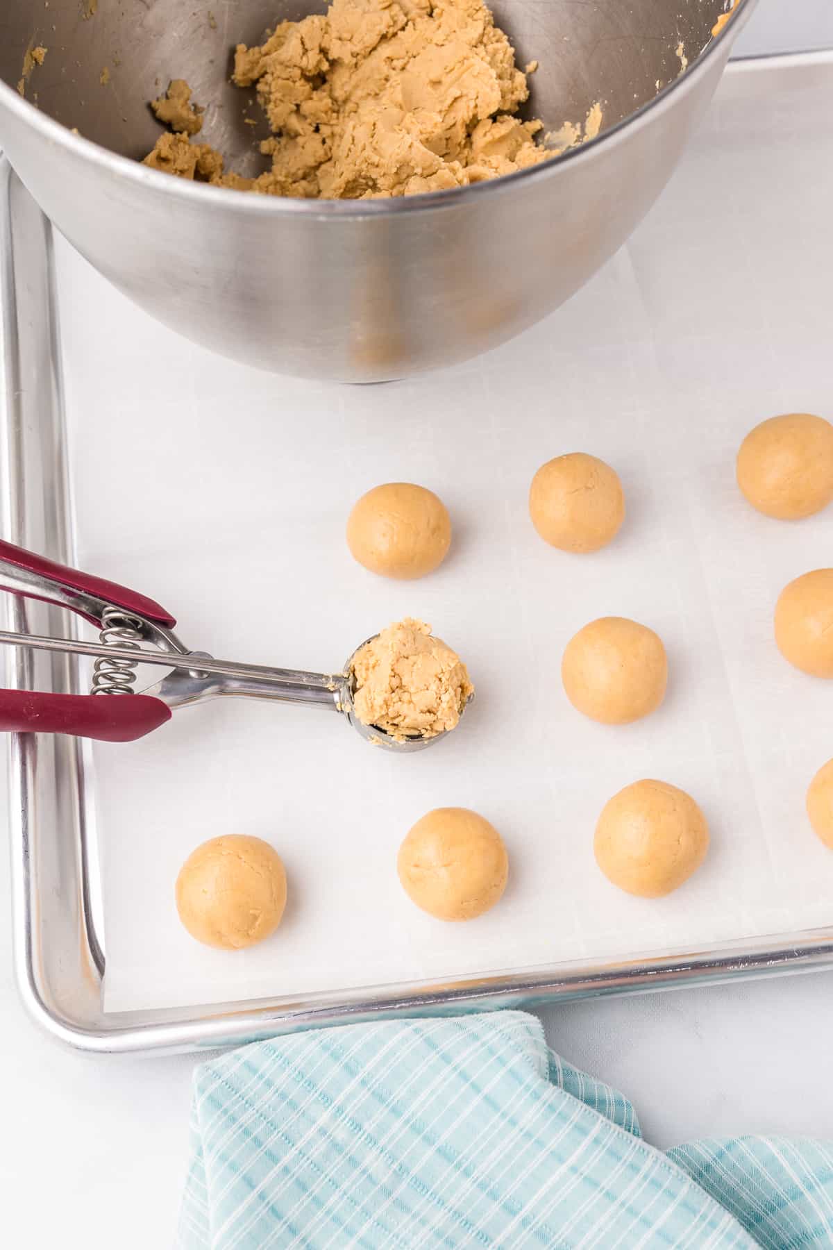 Rolled peanut butter dough on a sheet pan next to a small cookie scoop full of dough with a mixing bowl full of dough nearby.
