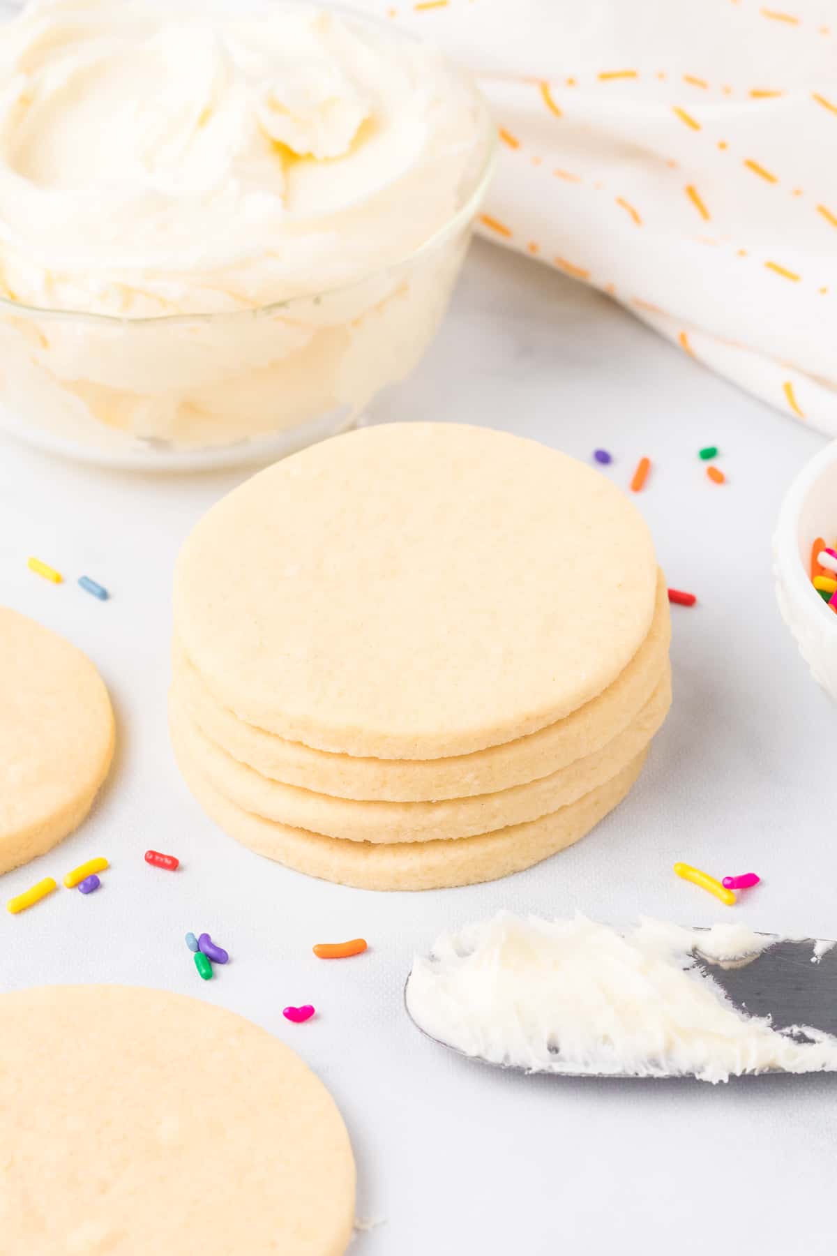 Stacked round sugar cookies nest to a bowl of frosting and a frosting covered knife on a counter from the side.