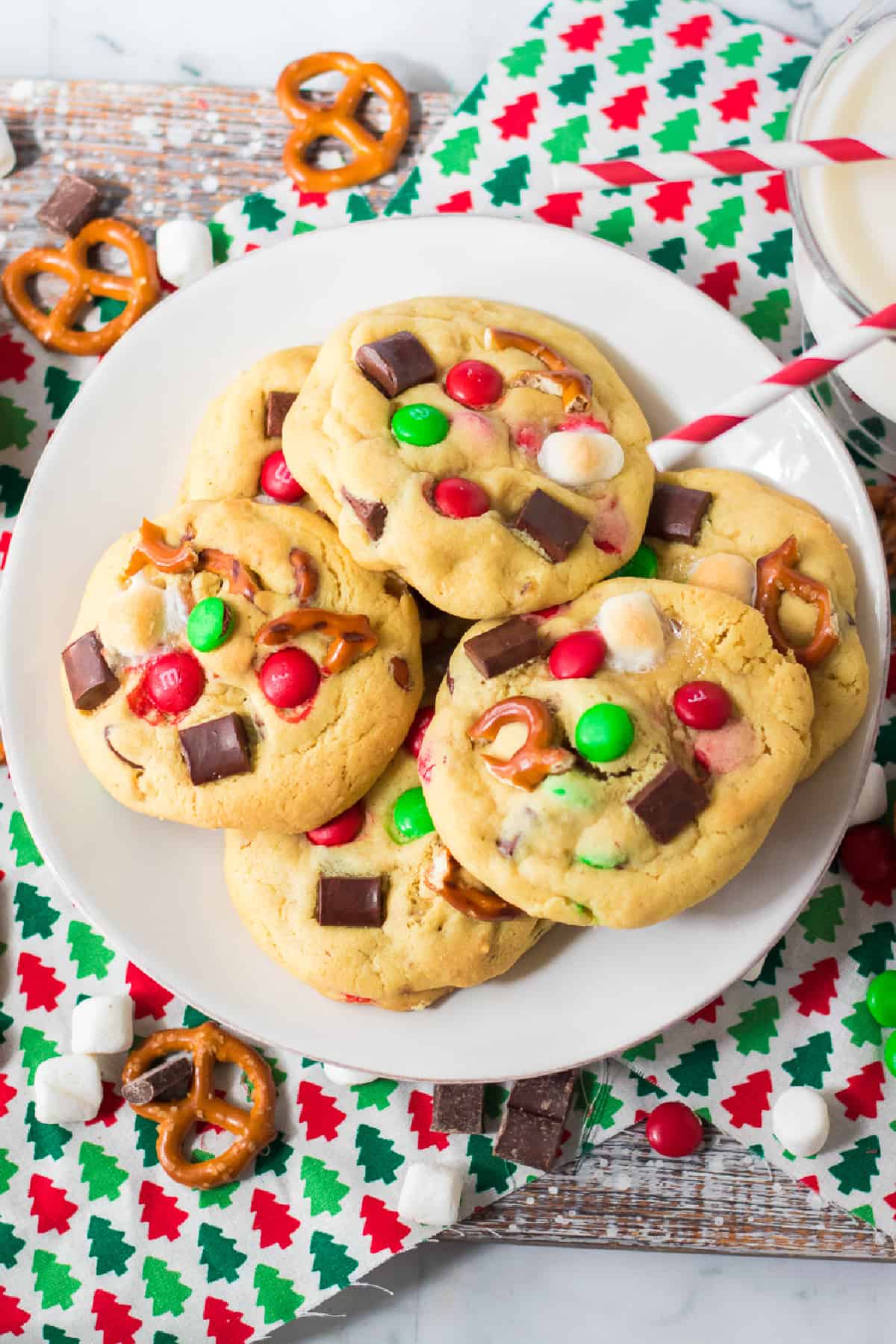 Plate of cookies covered in red and green M&Ms, chocolate chunks, marshmallows and pretzel pieces on a counter from above with a glass of milk nearby.