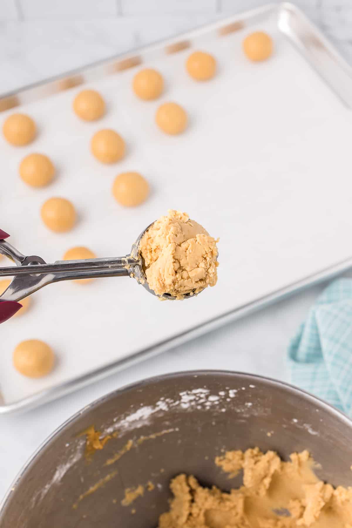 Scooping peanut butter dough with a cookie scoop from a large mixing bowl into portions on a baking sheet on a counter from the side.