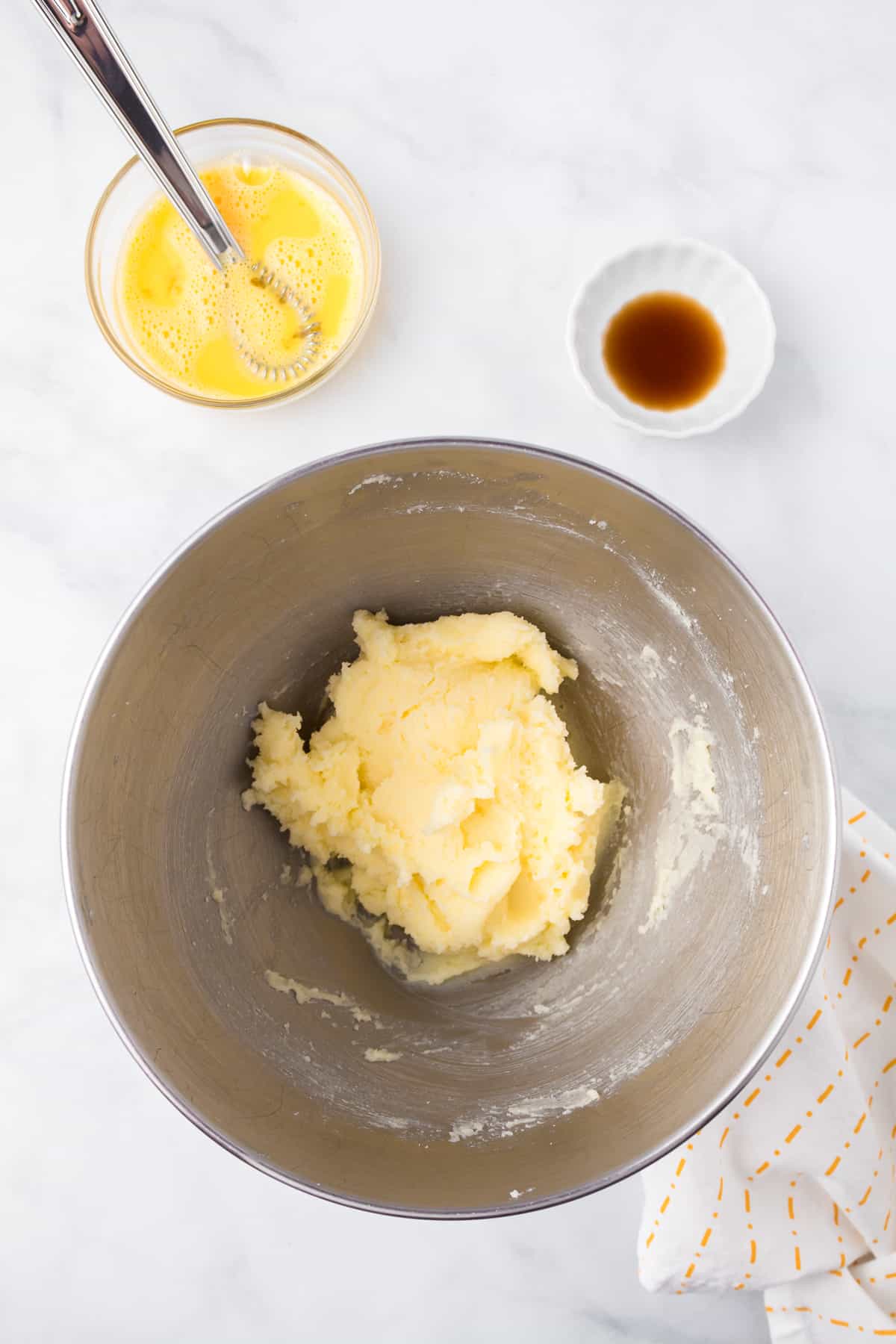 Creamed butter and sugar in a mixing bowl from overhead on a counter with a bowl full of beaten egg and a small bowl full of vanilla on a counter from overhead.