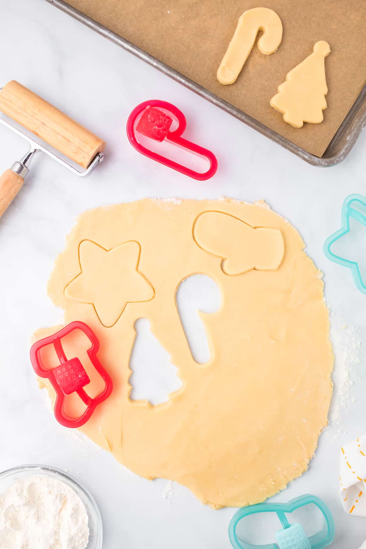 Sugar cookie dough rolled out on a counter from overhead being cut out with cookie cutters while a pan with some cookies, a bowl of flour, and a rolling pin sit nearby on the counter.
