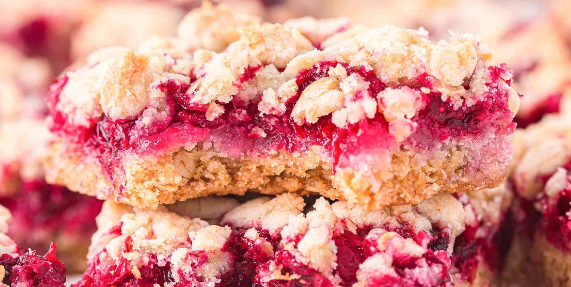 Wide close up view of a cranberry crumble bar stacked on top of a second bar missing a bite so you can see the fruity cranberry layer under the crumble.