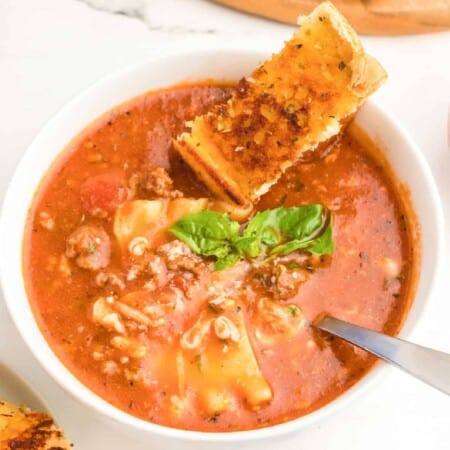 Lasagna soup in a bowl with a spoon topped with mozzarella cheese, basil and a slice of garlic bread.