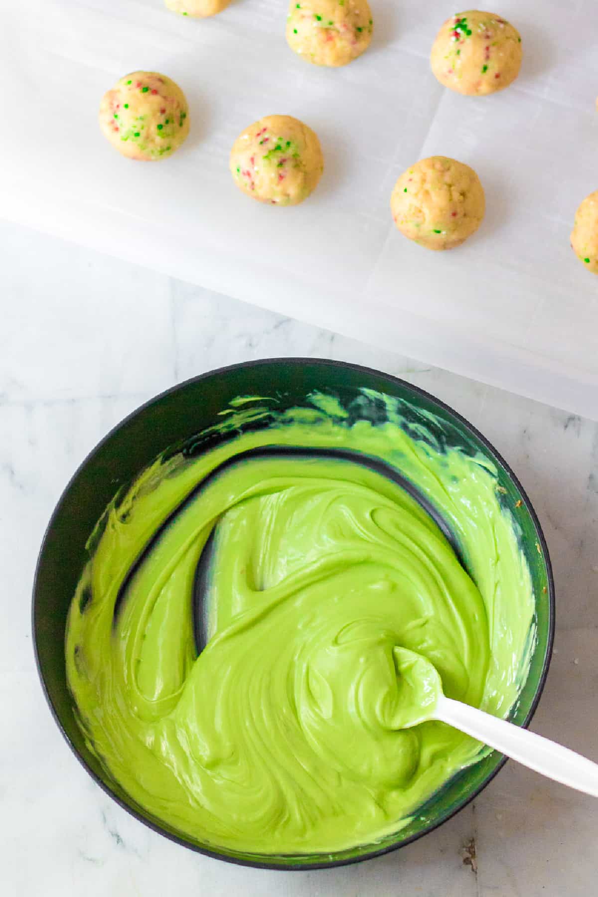 Bright green melting wafers melted in a bowl with a spoon withgolden Oreo truffles on parchment paper nearby.