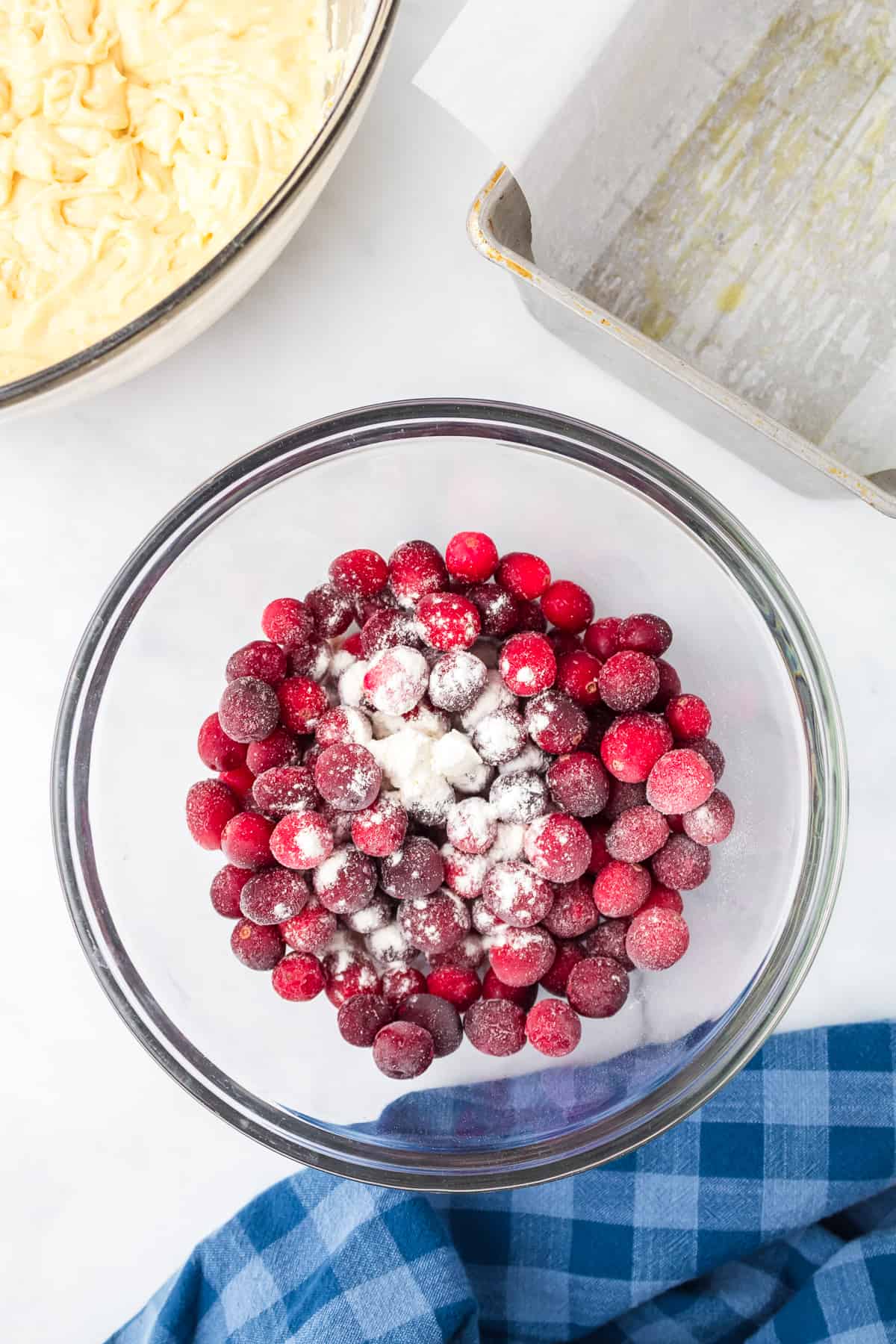 Cranberries being tossed with flour in a large mixing bowl with a second boil containing batter and a loaf pan lined with parchment paper nearby on the counter.