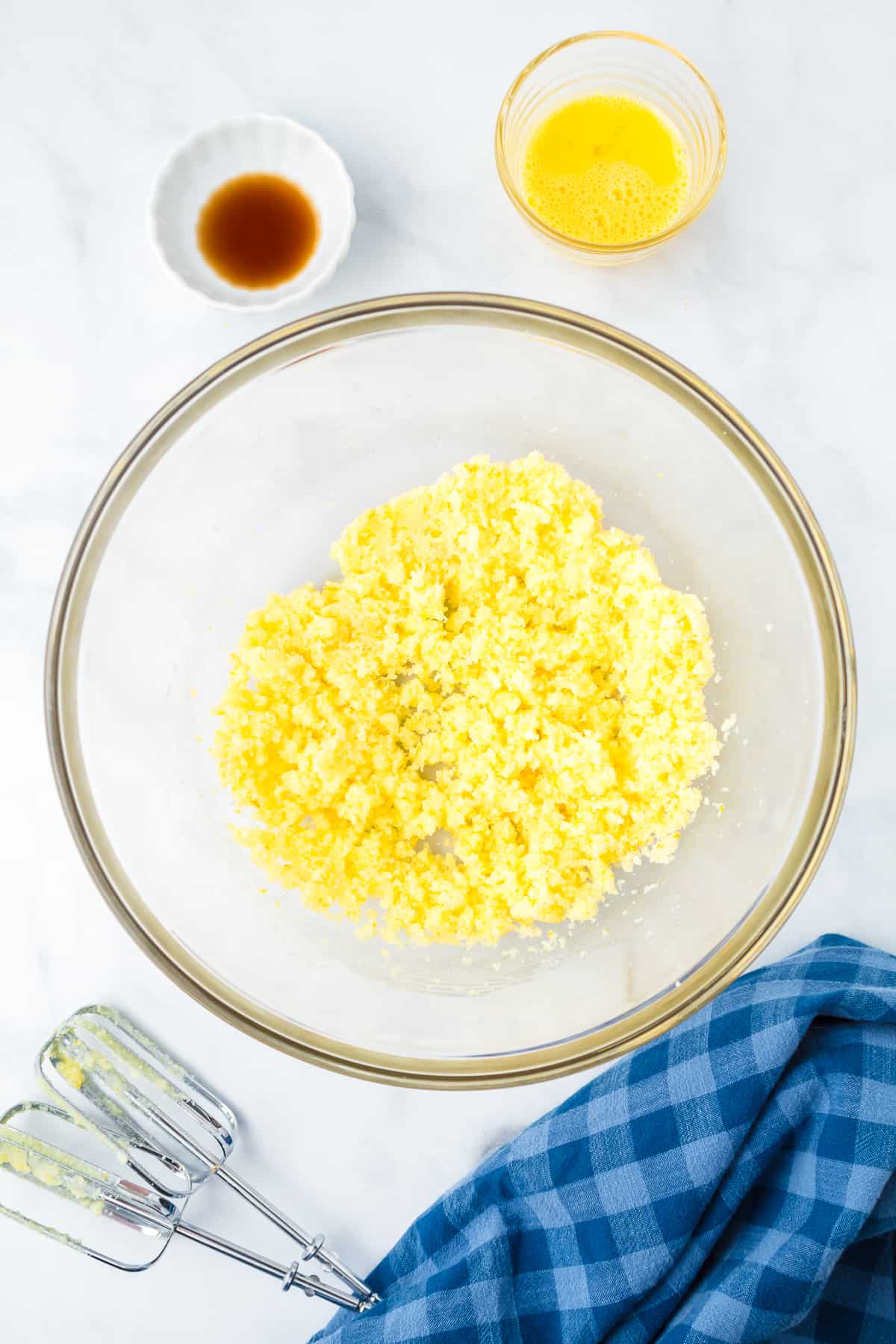 Butter and sugar creamed in a mixing bowl from above with vanilla and  beaten egg in small bowls nearby on the counter.