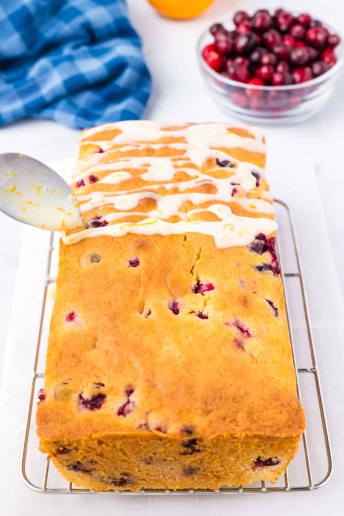 White colored glaze being drizzled on a loaf of cranberry orange bread on a wire rack with a spoon from the end of the loaf..