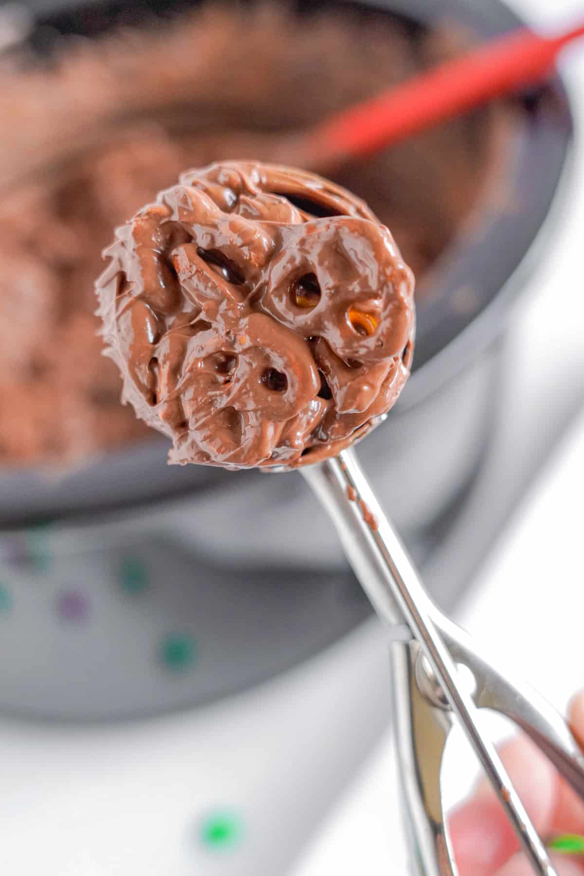 Scooping chocolate pretzel candy with a cookie scoop from a crockpot in the background.