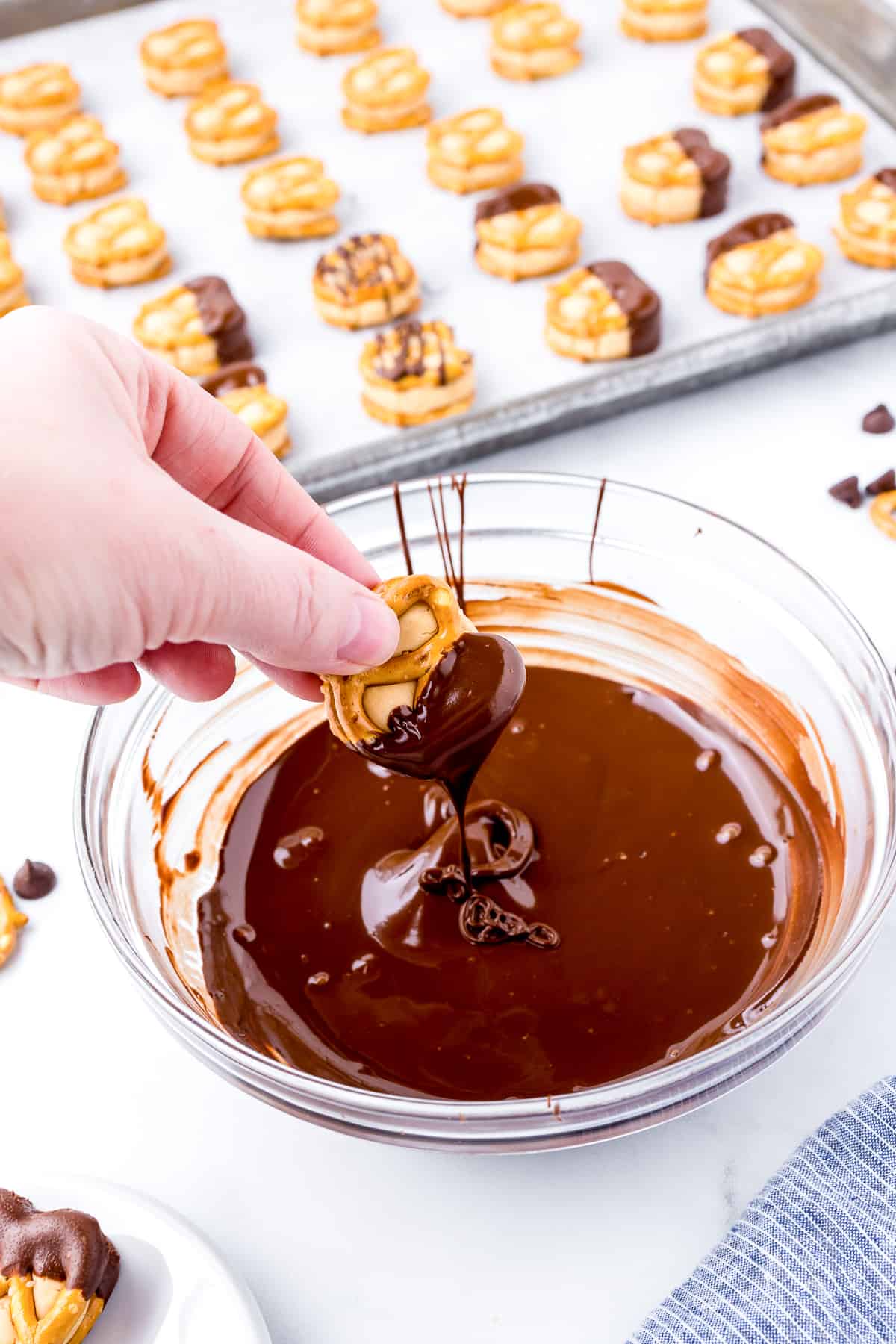 Peanut butter pretzel bite being dipped in chocolate in a bowl with more on a sheetpan in the background.