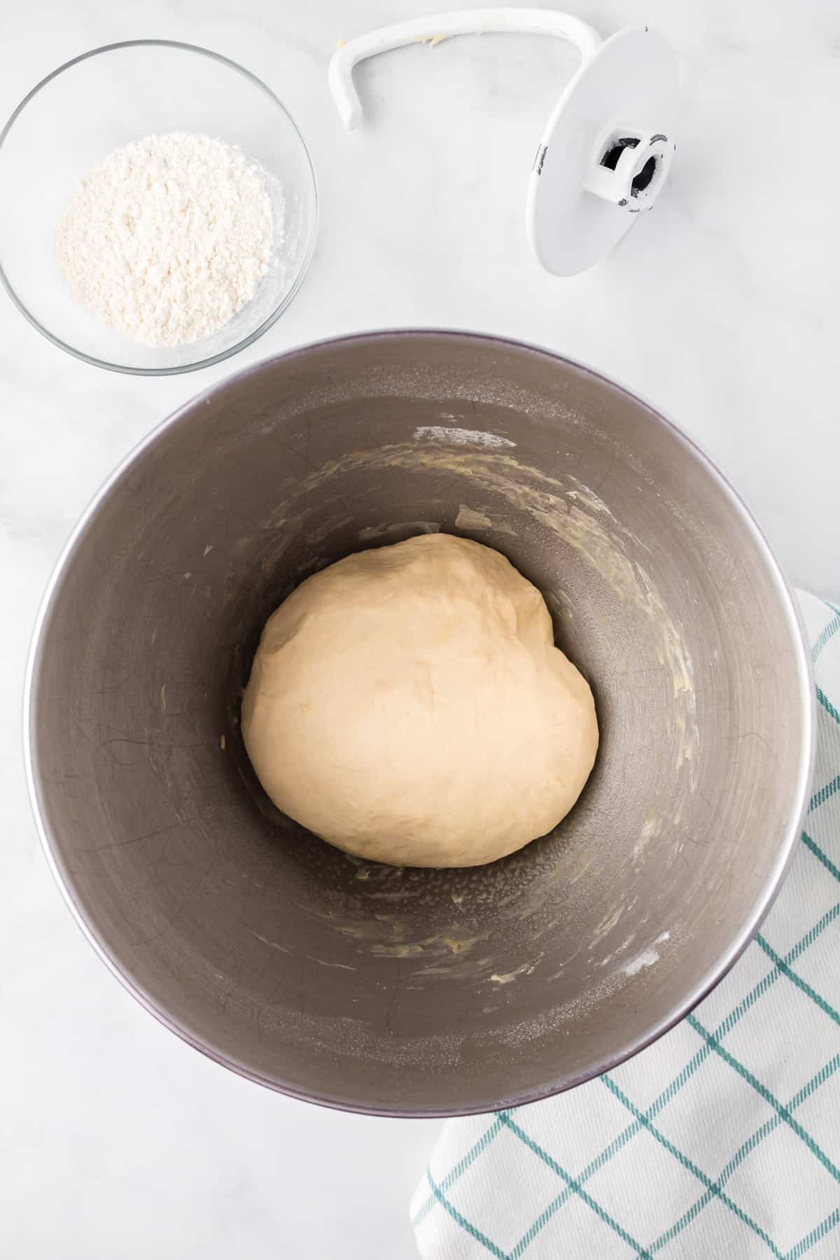 Garlic knot dough after mixing in a large mixing bowl with a dough hook and more flour in a bowl nearby on the counter from overhead.