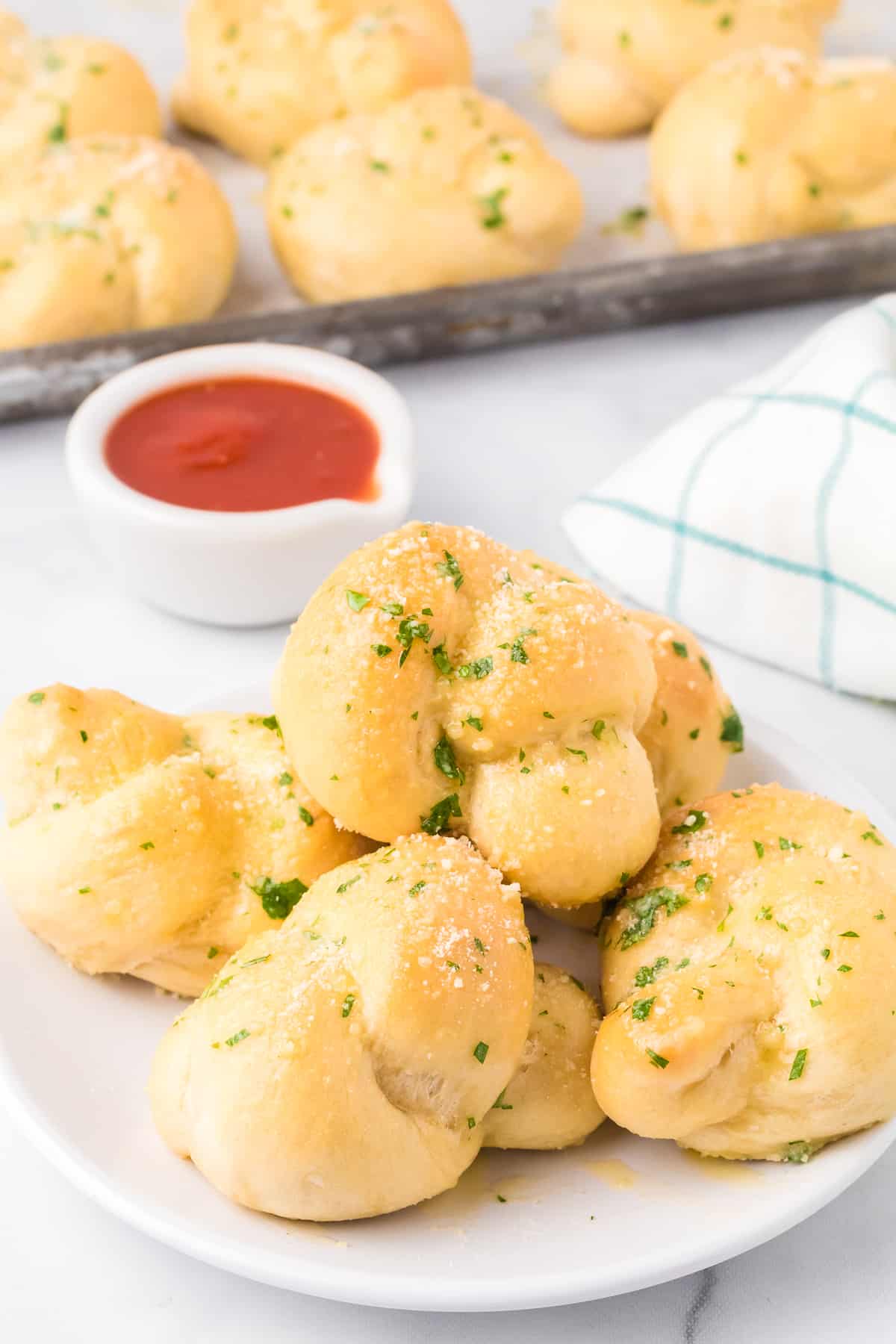 Garlic knots piled on a plate from the side on a counter covered in garlic, parsley and cheese.