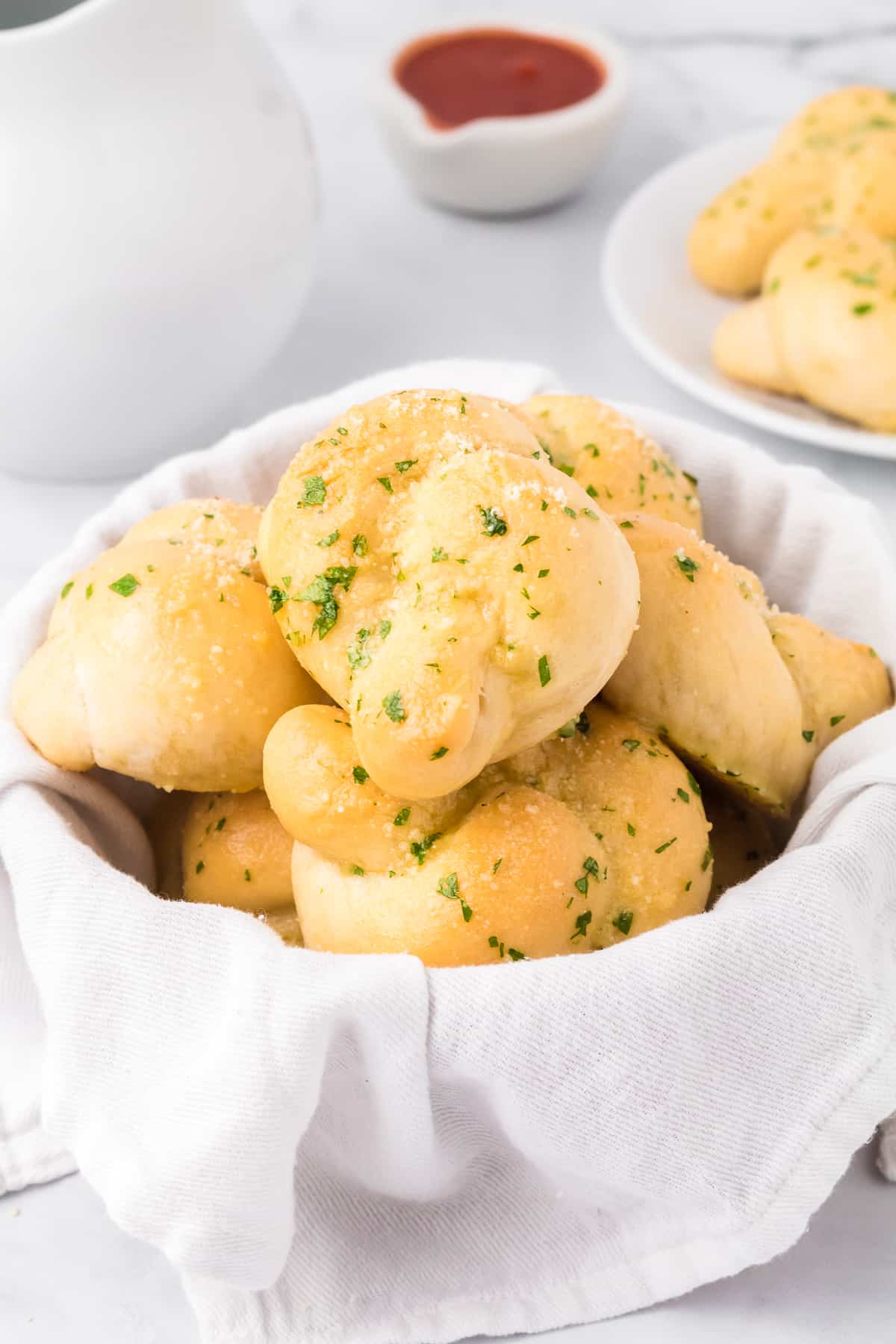 Garlic knots in a bread bowl with a small bowl of tomato sauce and a plate with more garlic knots in the background on a counter.