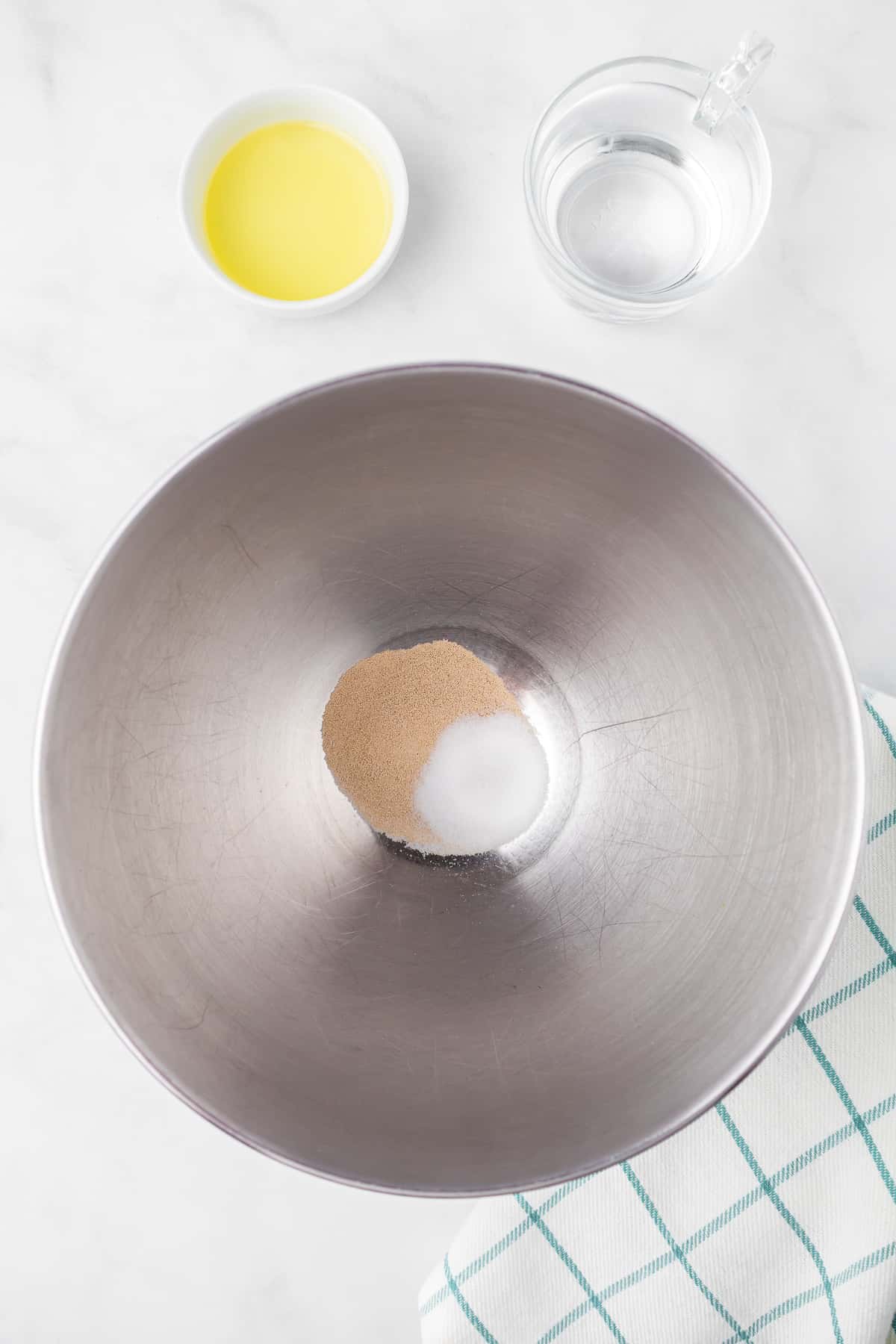 Yeast and sugar in a large mixing bowl from overhead with water and olive oil nearby in bowls on the counter.