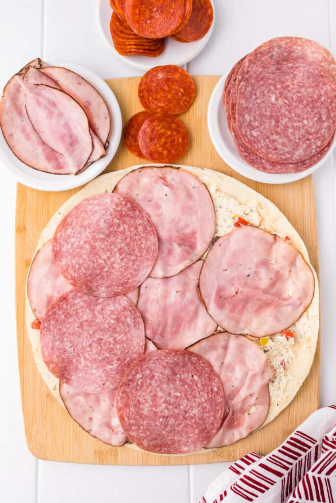 Adding ham and salami to the top of a large tortilla from overhead.