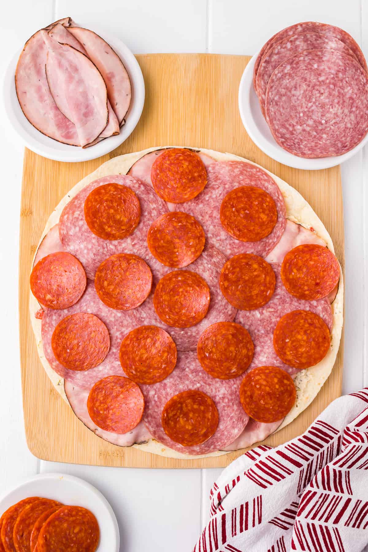 Adding pepperoni to the italian pinwheel tortilla from above on a cutting board with more Italian meats on plates nearby.