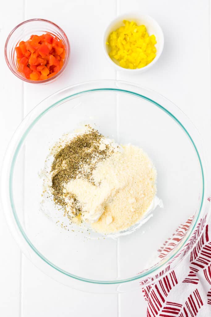 Mixing cream cheese with seasoning in a large bowl from overhead with diced roasted pepper and pepperoncini nearby in bowls on the counter.