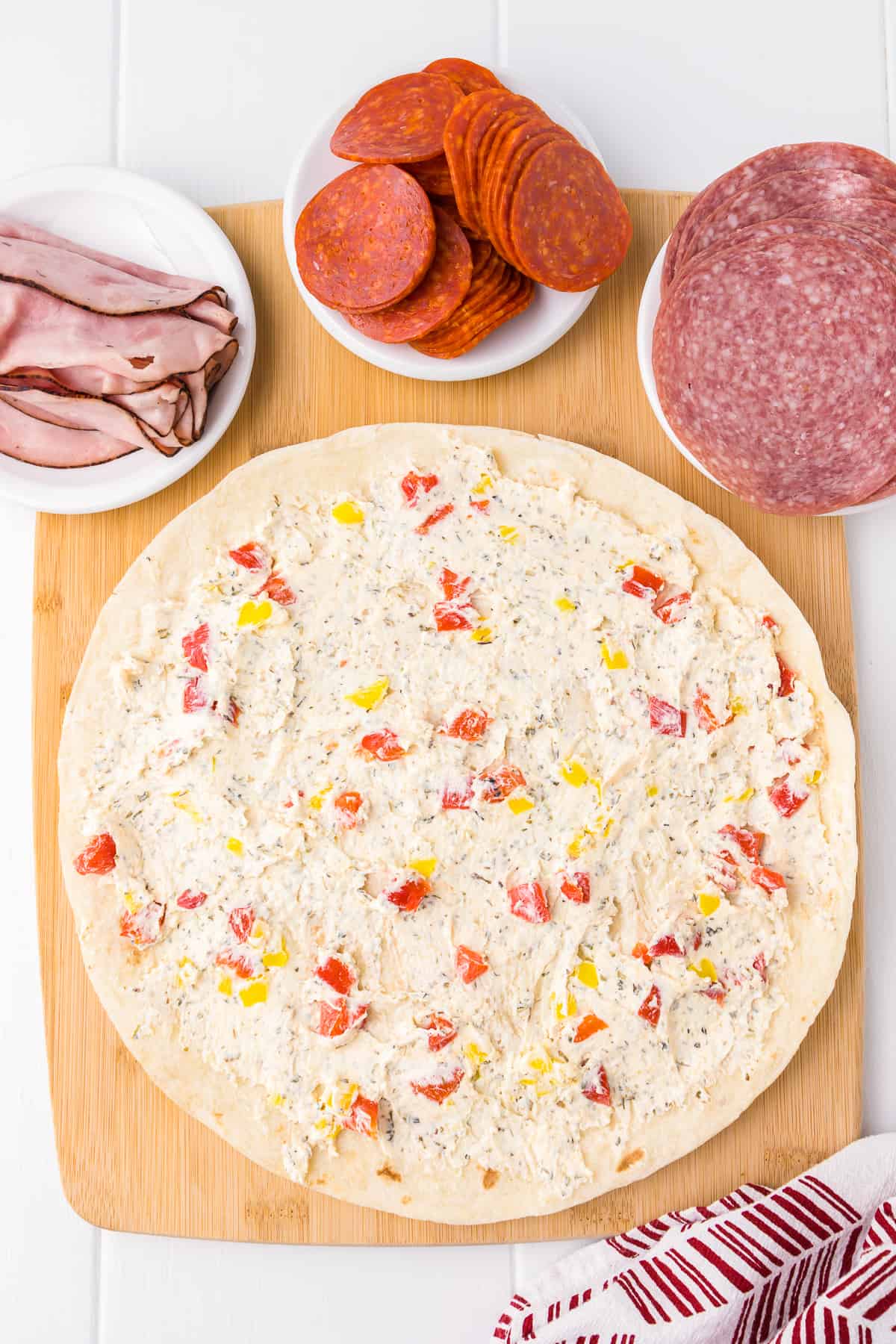 Spreading the cream cheese mixture full of diced peppers on a tortilla from overhead on a cutting board with ham, pepperoni and salami on small plates nearby.