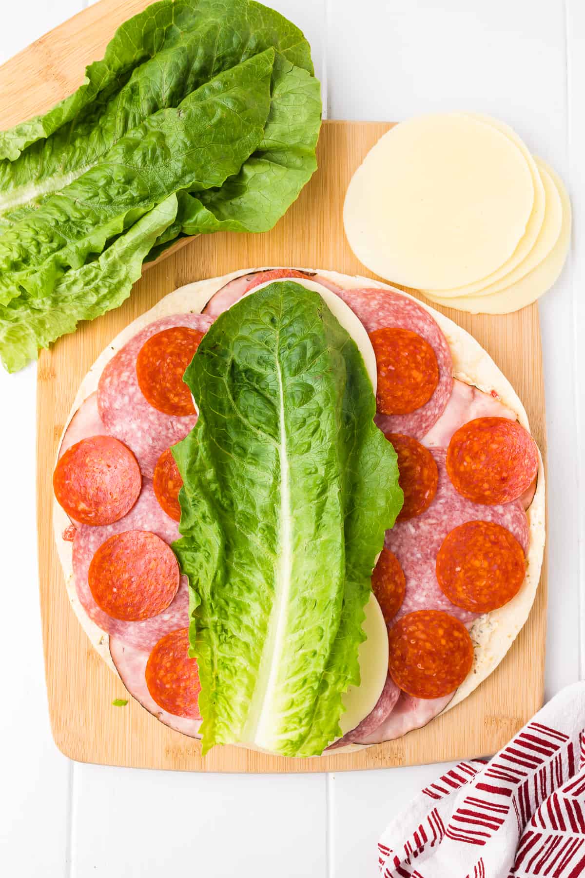 Adding lettuce to the top of a tortilla covered in Italian meats and cheese.