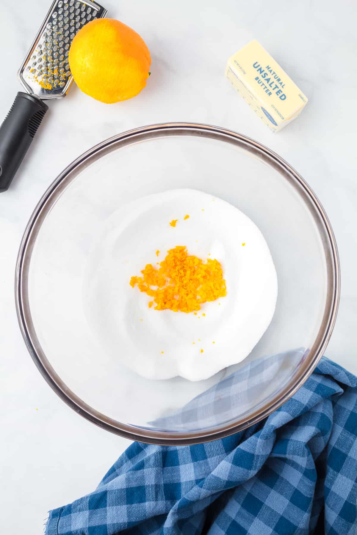Orange zest being mixed into sugar in a bowl from above with a zester and an orange nearby on the counter.