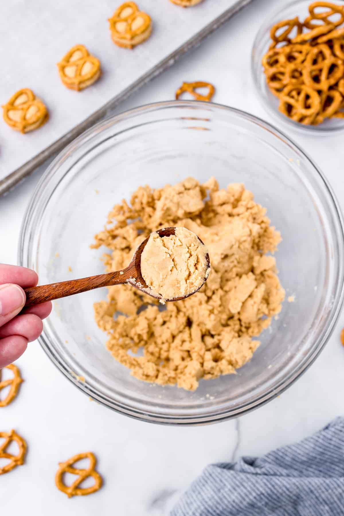 Scooping peanut butter pretzel dough from a bowl below with a cookie sheet with more peanut butter dough balls nearby.