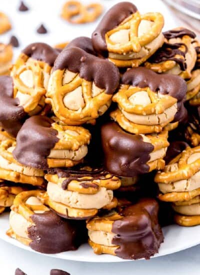Peanut butter stuck between small pretzels dipped in chocolate stacked high on a plate from the side on a counter.