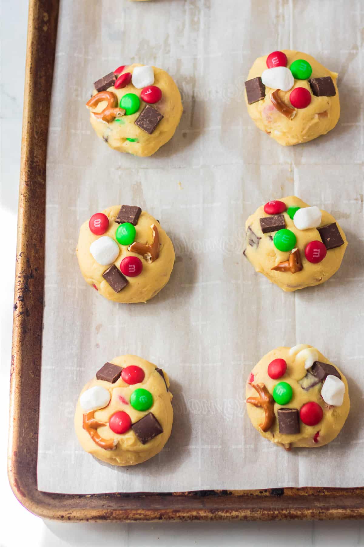 Baking sheet with cookie dough balls covered in chocolate chunks, red and green M&Ms, marshmallows and pretzels pieces.