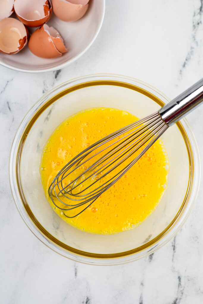 Eggs scrambled in a bowl with a whisk and cracked egg shells nearby.