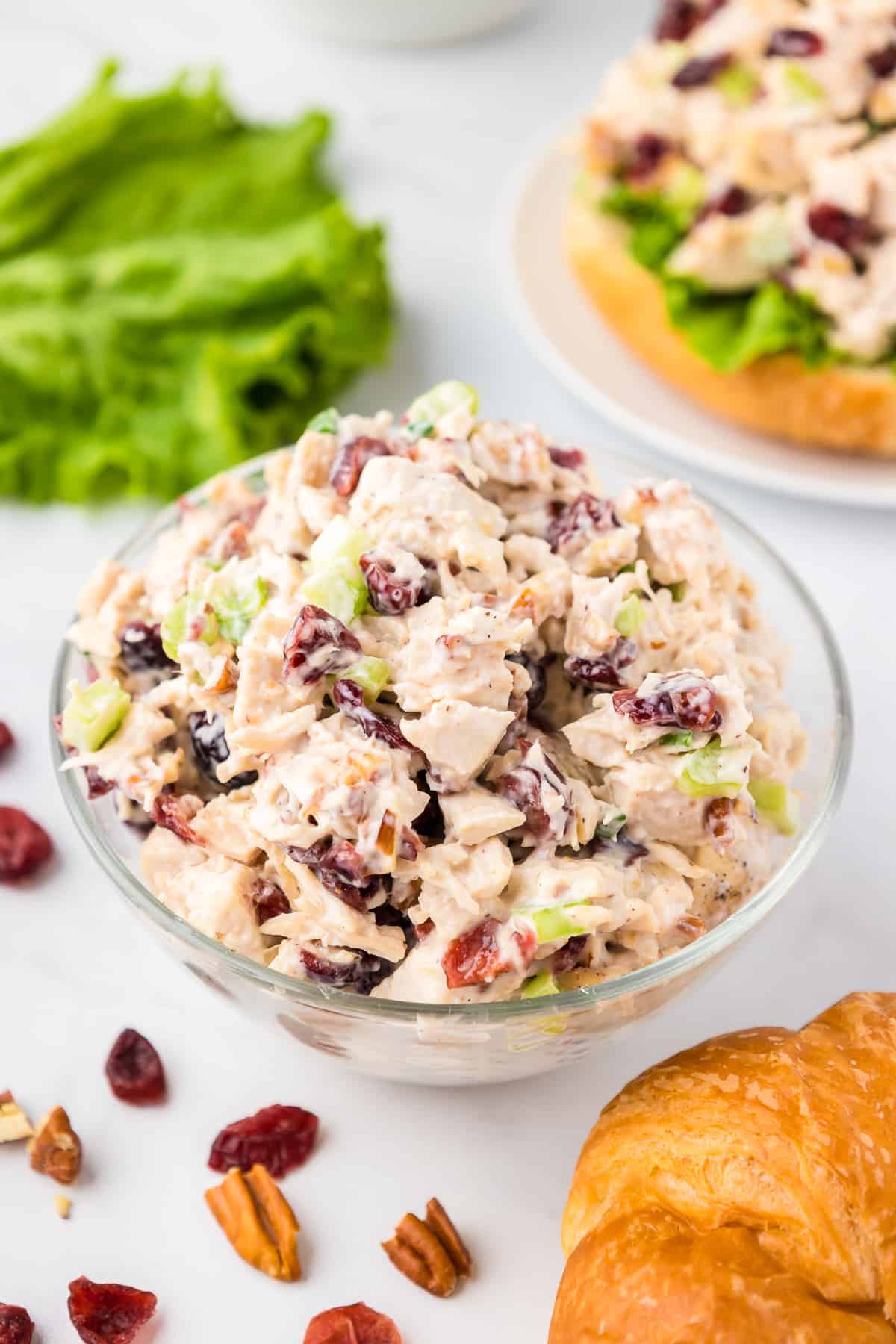 Cranberry pecan chicken salad mixed in a bowl on a counter with lettuce, croissants and part of a sandwich on the counter.