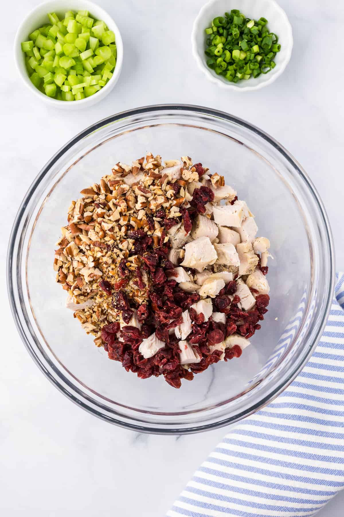 Diced chicken being mixed with dried cranberries and pecans in a large bowl from overhead on a counter with celery and green onion nearby.