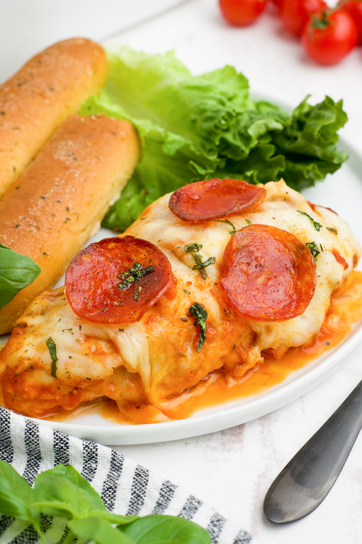 Stuffed pizza chicken breast on a plate covered in cheese and pepperoni next to breadsticks and salad from the side.