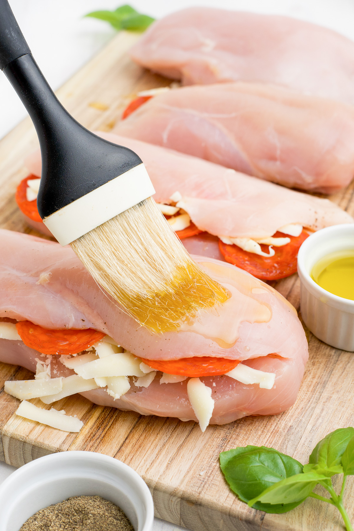 Raw chicken breasts stuffed with cheese and pepperoni being brushed with oil from the side on a cutting board.