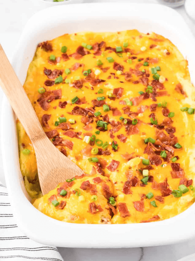Side view of a pan of loaded mashed potato casserole on a counter topped with cheese and bacon being scooped with a wooden spoon.