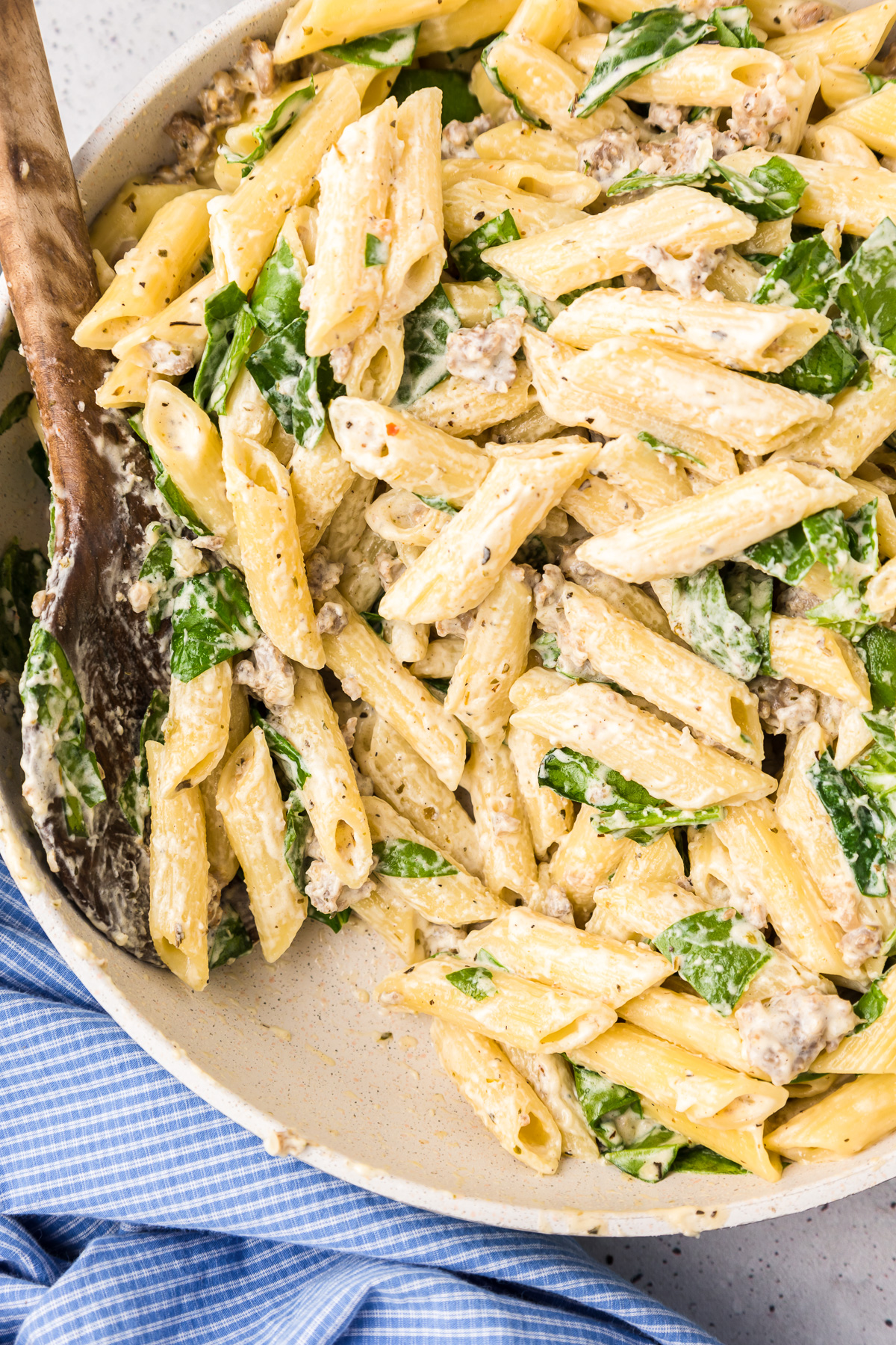 Creamy penne pasta in a white sauce with sausage and spinach in a pan from the side with a large wooden spoon in the pan.