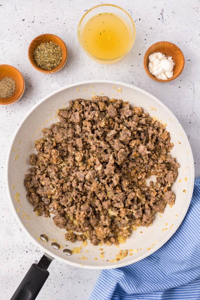 Loose sausage browning in a skillet from overhead with spices, chicken broth and flour in small bowls nearby.