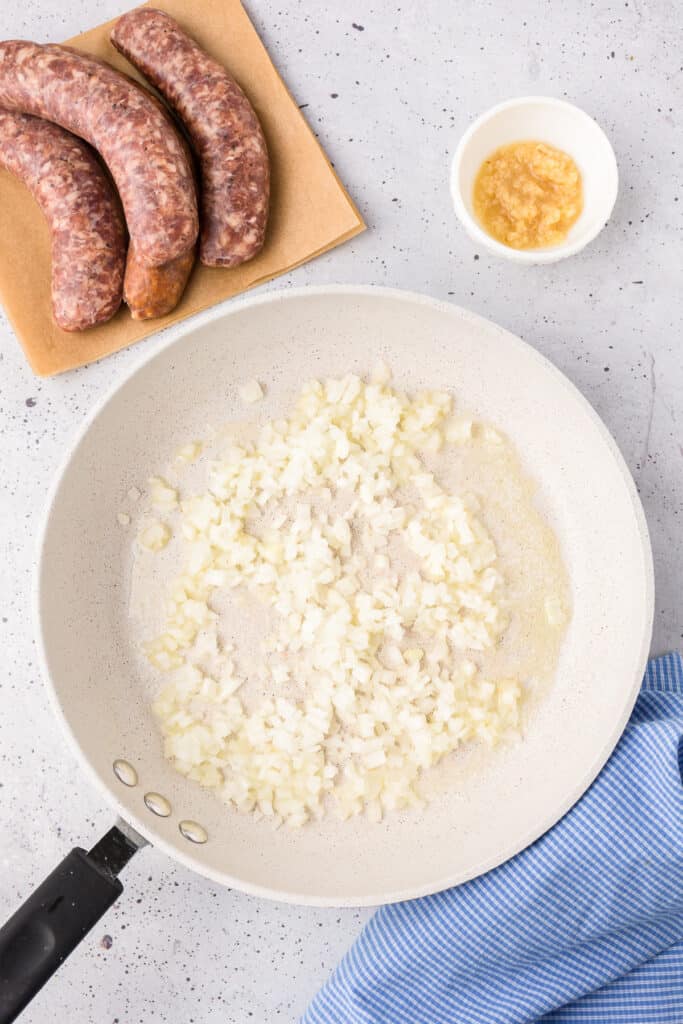 Cooking onion in a large skillet from overhead with raw sausage links on a cutting board and garlic in a small bowl nearby.