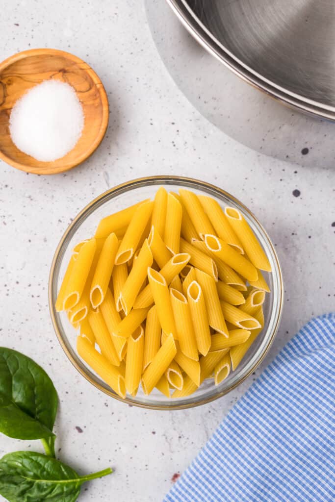 Penne pasta in a large bowl next to a pot of water and a small bowl of salt from overhead.