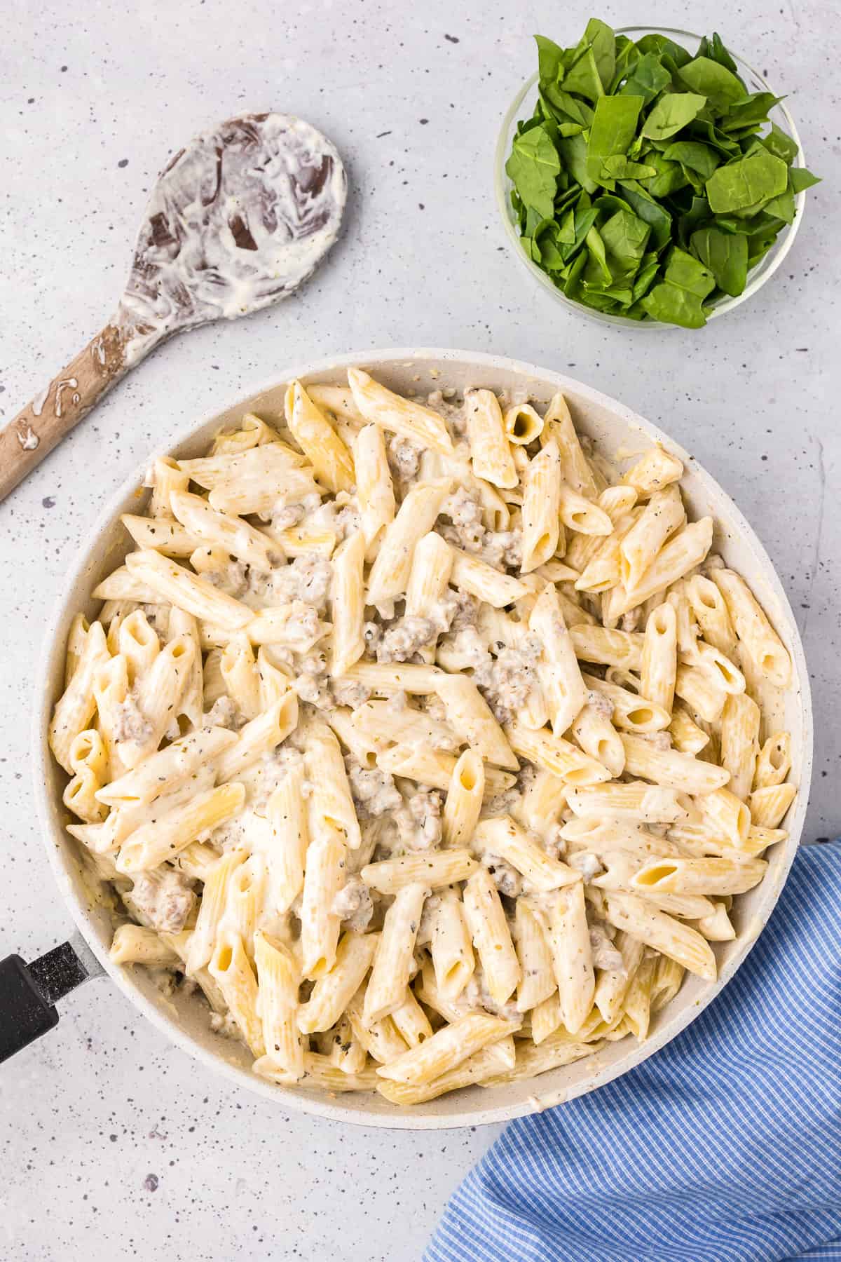 Pasta being mixed into the creamy parmesan cheese sauce in a large skillet with a bowl of chopped spinach and a large spoon covered in cheese sauce nearby from above.