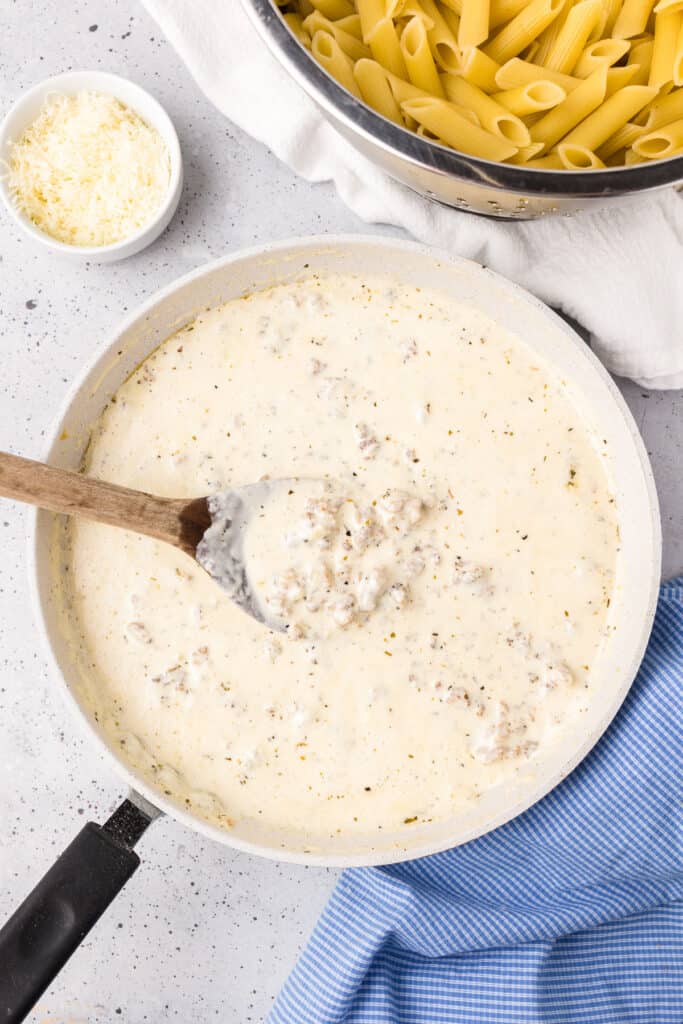 Creamy sauce full of sausage pieces in a large skillet being stirred with a large spoon from above with pasta and parmesan cheese nearby .