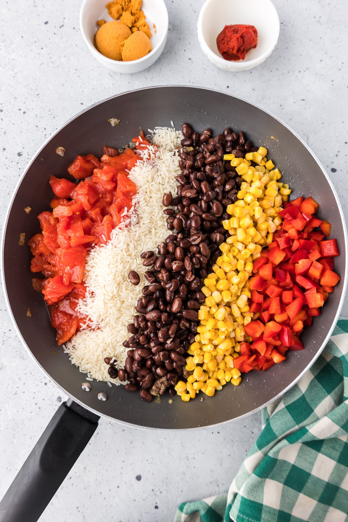 Tomatoes, rice, black beans, corn and red bell pepper added in rows in a skillet to cook with tomato paste and taco seasoning in small bowls nearby.