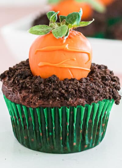Close up of a cupcake with an orange chocolate covered strawberry decorated to look like a carrot in chocolate cookie crumble dirt.