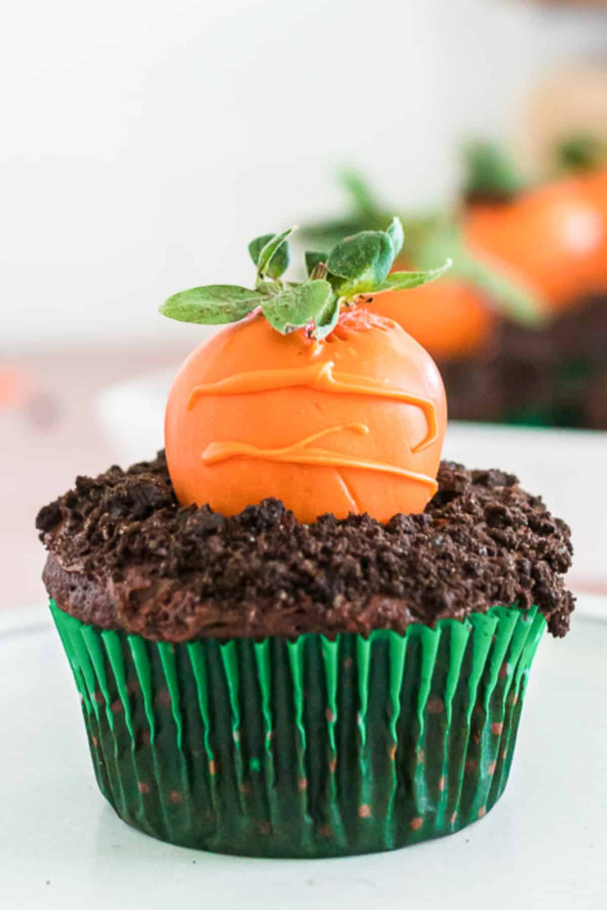 Close up of a cupcake decorated to look like a carrot in the ground with chocolate and an orange chocolate dipped strawberry on top close up from the side.