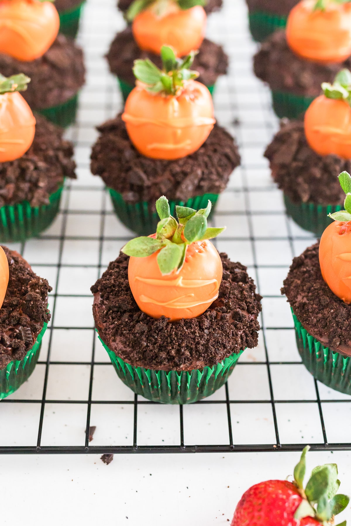 Close up of chocolate cupcakes that look like dirt with chocolate cookie crumbs topped with strawberries covered in orange chocolate to look like carrots.