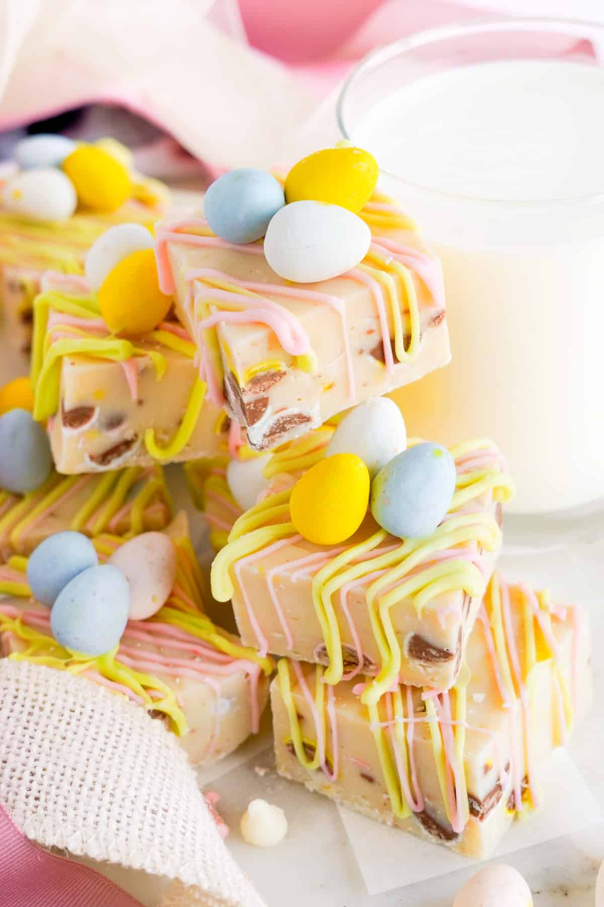 Multiple pieces of easter fudge decorated with mini chocolate eggs stacked high next to a glass of milk.