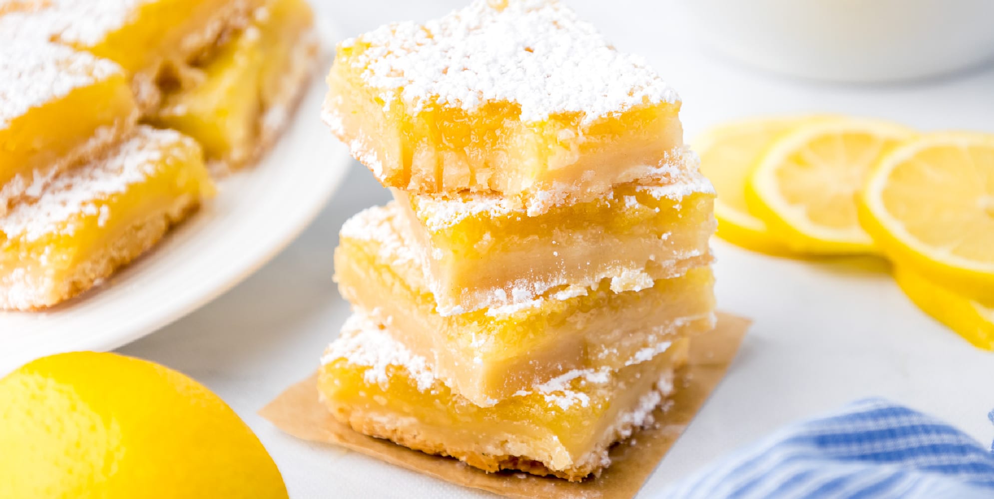 A wide view of lemon bars stacked high with the top bar missing a bite, and more bars and lemons in the background.