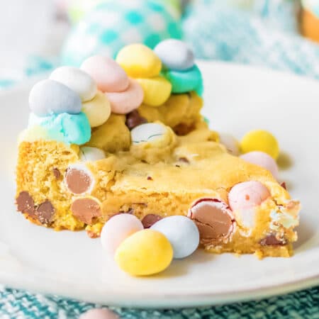 Slice of chocolate mini egg cookie cake on a plate decorated with more candy Easter eggs from the side.