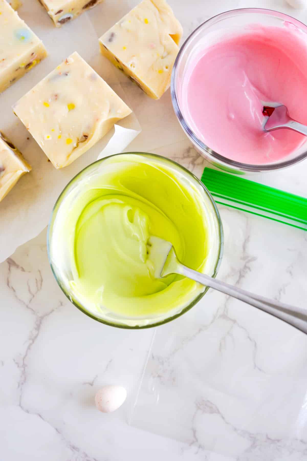 Green and pink candy melts melted in bowls with a fork to stir from above next to pieces of white chocolate fudge.